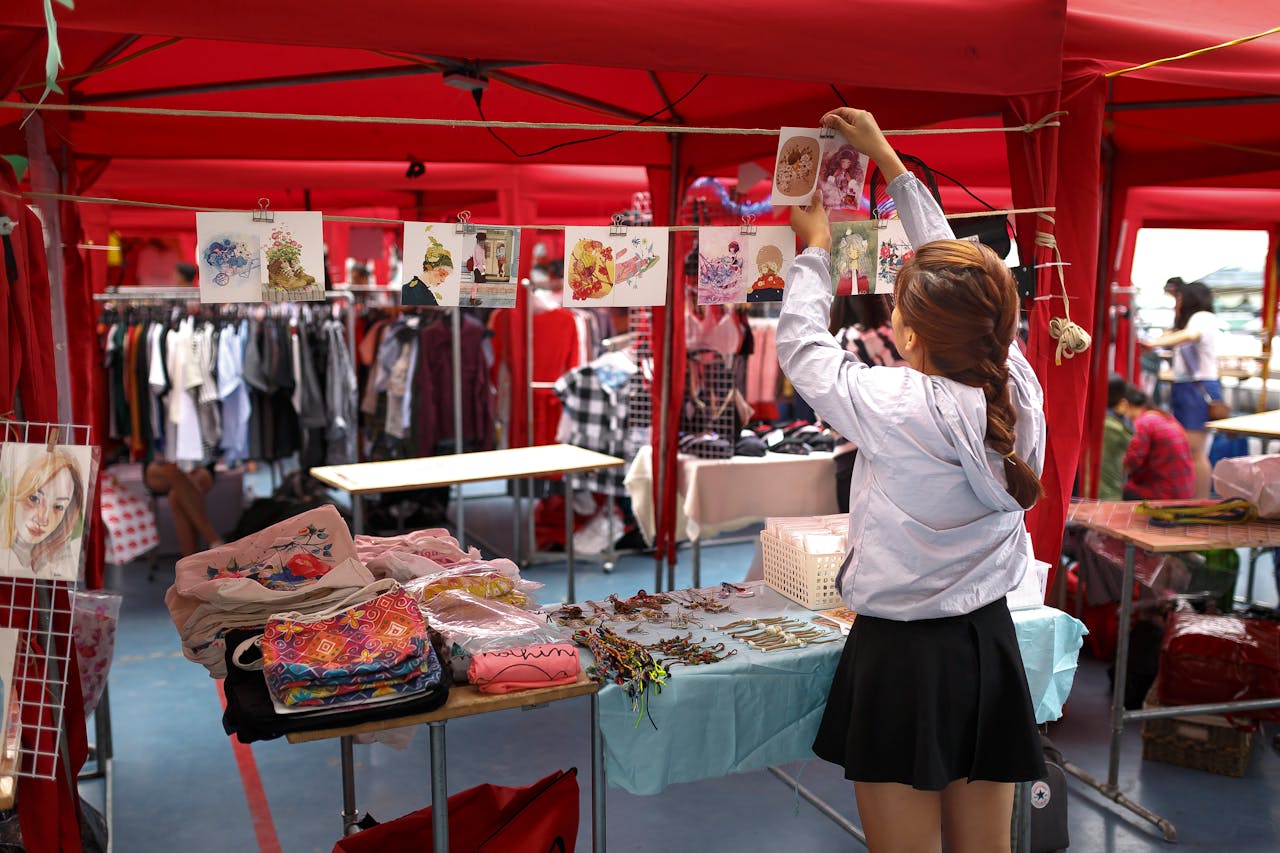 Home A woman organizes art prints in a vibrant outdoor market stall with colorful clothing and accessories.