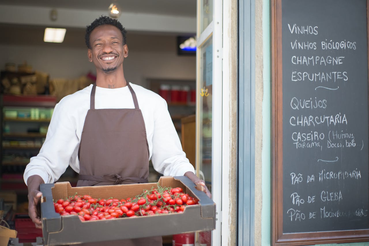 About Smiling vendor standing outside a Portuguese store holding a box of ripe tomatoes. Perfect for grocery or food themes.